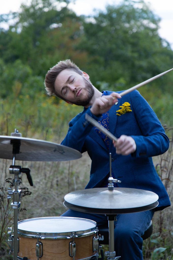 Lee Fish playing the drums in a field. Trees are visible in the background.